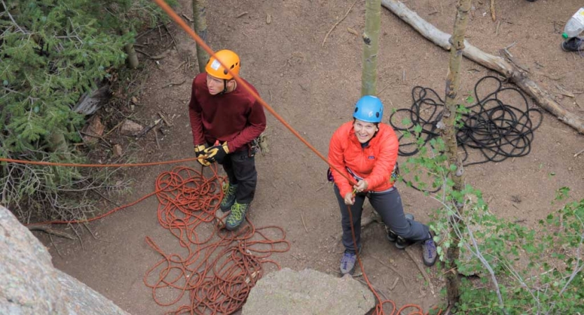 From above, two people wearing safety gear look up as they belay unseen climbers. 